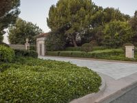the gate to the private home is located behind a garden of shrubs and trees with concrete pillars
