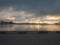 a lone bench sits on the bank of a lake during the sunset with the city in the background