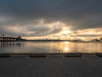 a lone bench sits on the bank of a lake during the sunset with the city in the background