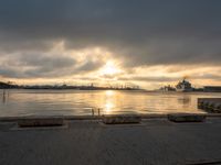 a lone bench sits on the bank of a lake during the sunset with the city in the background