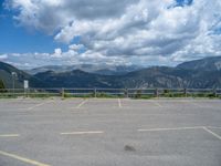 a car park with mountains and a chain link fence behind it in the background are clouds and a body of water