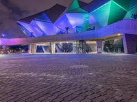 an illuminated up building surrounded by stone pavement and lights on in the distance is people