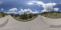 a street view from the perspective angle of an intersection of a road, and on it there is a forest and mountains with clouds