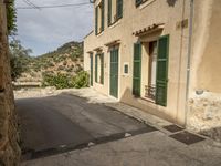 a narrow, paved street in a deserted town with stone walls and green shuttered doors