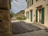 a narrow, paved street in a deserted town with stone walls and green shuttered doors