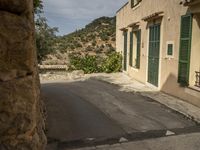 a narrow, paved street in a deserted town with stone walls and green shuttered doors