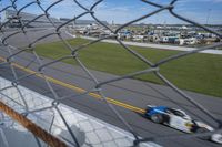 race cars race up an empty track as seen from behind a chain - link fence