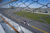 race cars race up an empty track as seen from behind a chain - link fence