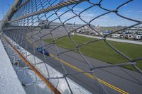 race cars race up an empty track as seen from behind a chain - link fence