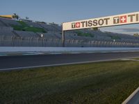 a long race track with a sign and racing cars on it at dusk with stadium bleachers in the background