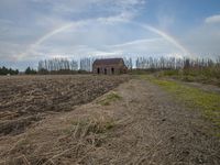 a rainbow is shining in the sky above an empty field in front of a house and a small structure in the middle of the distance