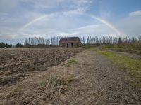 a rainbow is shining in the sky above an empty field in front of a house and a small structure in the middle of the distance