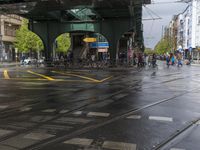 a rainy intersection with people walking around on the street and buildings in the background and people standing underneath it