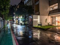 a rainy sidewalk at night with green plants in front and some people sitting on a bench