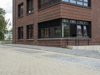 brick sidewalk on side of building with large windows and plants outside, and sidewalk blocker along street