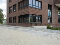 brick sidewalk on side of building with large windows and plants outside, and sidewalk blocker along street