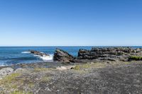 some rocks and water on the shore line the beach under a blue sky with some green grass