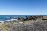 some rocks and water on the shore line the beach under a blue sky with some green grass