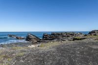 some rocks and water on the shore line the beach under a blue sky with some green grass