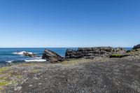 some rocks and water on the shore line the beach under a blue sky with some green grass