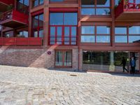 the view of the outside of a building with windows on all three floors, and people walking on stone pavement