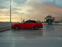 red car parked in an empty parking lot with a cloudy sky behind it, surrounded by a building