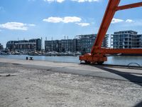 a red crane sitting on the sidewalk next to the water and buildings behind it, with blue sky in background