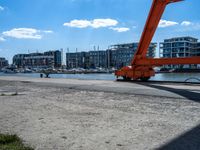 a red crane sitting on the sidewalk next to the water and buildings behind it, with blue sky in background