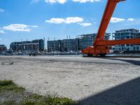 a red crane sitting on the sidewalk next to the water and buildings behind it, with blue sky in background