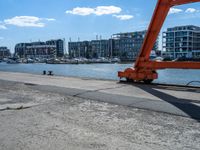 a red crane sitting on the sidewalk next to the water and buildings behind it, with blue sky in background