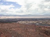 a red dirt hill with a dog sitting on the edge, as well as water, mountains and some trees