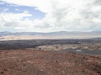 a red dirt hill with a dog sitting on the edge, as well as water, mountains and some trees