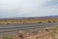 an empty highway in a flat desert land near mountains and grass, with some yellow wildflowers