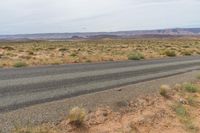 an empty highway in a flat desert land near mountains and grass, with some yellow wildflowers