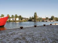 red ship docked on brick walkway next to body of water with city skyline and buildings