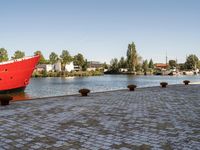 red ship docked on brick walkway next to body of water with city skyline and buildings