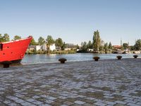 red ship docked on brick walkway next to body of water with city skyline and buildings