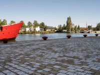 red ship docked on brick walkway next to body of water with city skyline and buildings