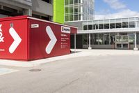 a red shipping container is at an intersection with another building and a street sign near it