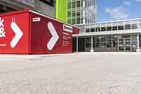 a red shipping container is at an intersection with another building and a street sign near it