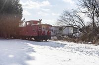 a red train car on tracks in the snow in front of houses and trees with trees