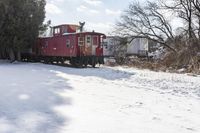 a red train car on tracks in the snow in front of houses and trees with trees