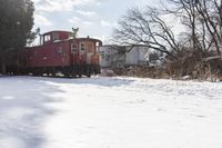 a red train car on tracks in the snow in front of houses and trees with trees