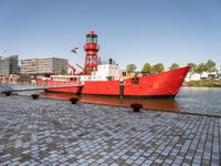 a red tug boat with a light tower in the background of a city and water