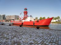 a red tug boat with a light tower in the background of a city and water