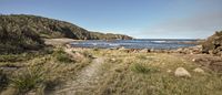 a wide beach with grass near a hill with rocks in the water and trees on one side