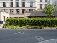 an empty street with bushes, benches and a building in the background - - - -