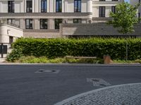 an empty street with bushes, benches and a building in the background - - - -