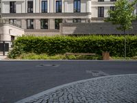 an empty street with bushes, benches and a building in the background - - - -