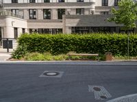 an empty street with bushes, benches and a building in the background - - - -